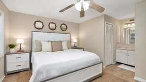 A bedroom with a large bed, bedside tables, a ceiling fan, and a bathroom with a vanity and mirror. - Newly furnished, Community Pool, Steps to Pier! (Saint Simons Island)