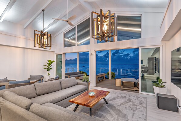 Enjoy expansive ocean views from the high ceiling and large windows in this spacious living area, featuring a comfortable sofa and contemporary chandelier.
