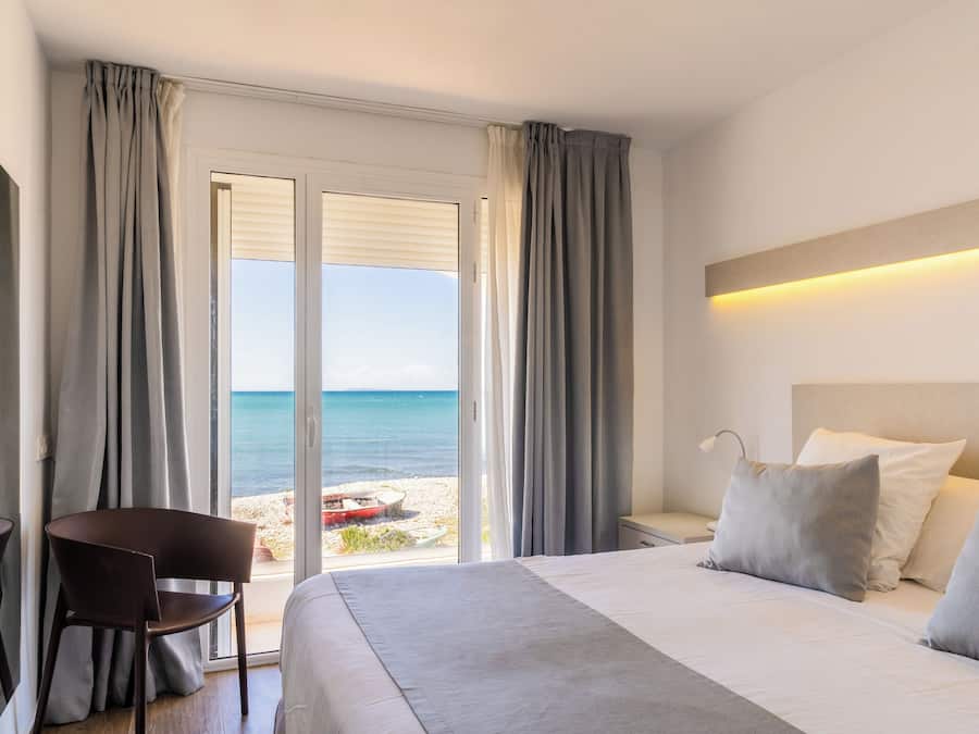 Habitación de hotel con cama, una silla, una mesita de noche y vista a la playa a través de una puerta corrediza.