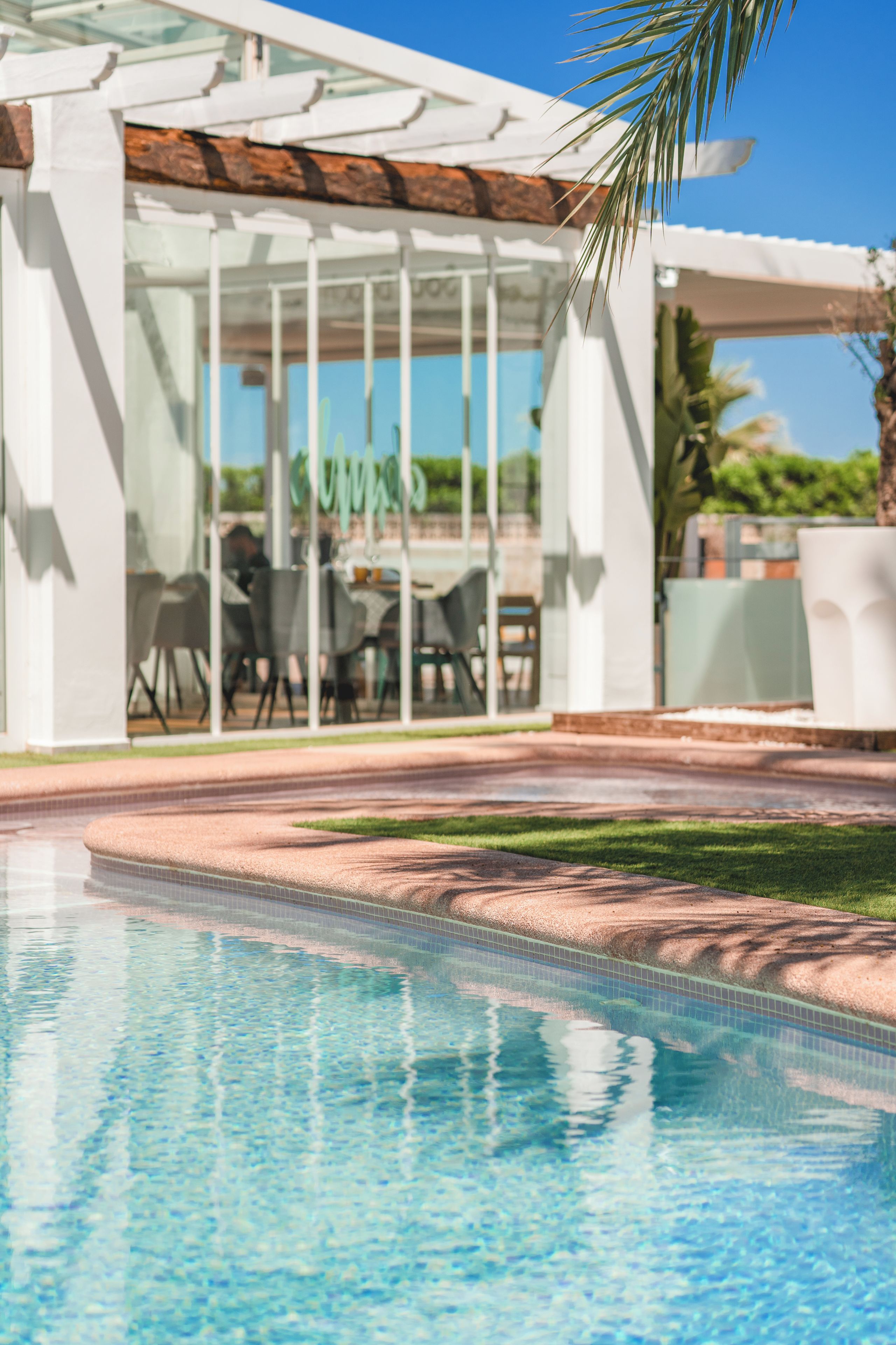 A swimming pool with a patio area, clear glass walls, and outdoor furniture.