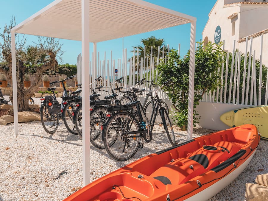 A covered area with bicycles parked under a white structure, a small orange boat, and a building with a white facade and a sign.