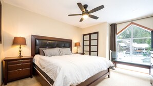 Ein Schlafzimmer mit einem großen Bett, zwei Nachttischen mit Lampen, einem Deckenventilator, einem Fenster mit Aussicht und einer Tür mit Milchglasverzierungen.