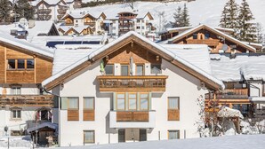 A snow-covered building with a balcony, wooden windows, and a ski slope in the background.