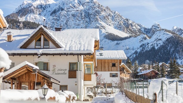 A snow-covered mountain lodge with wooden architecture, a visible sign reading 'casa Prima', and a clear view of the surrounding snowy mountains.