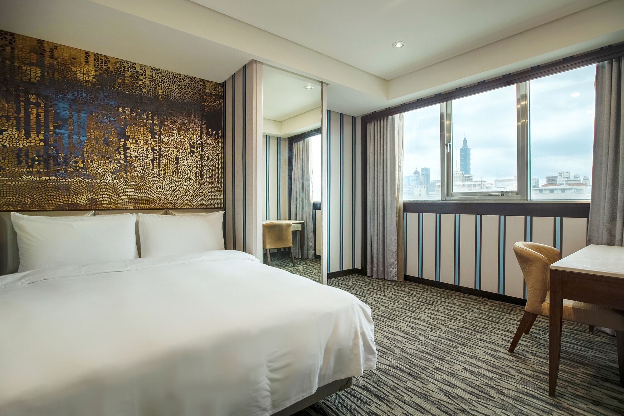 A hotel room with a large bed, a desk, and a chair. There is a view of a cityscape through large windows.