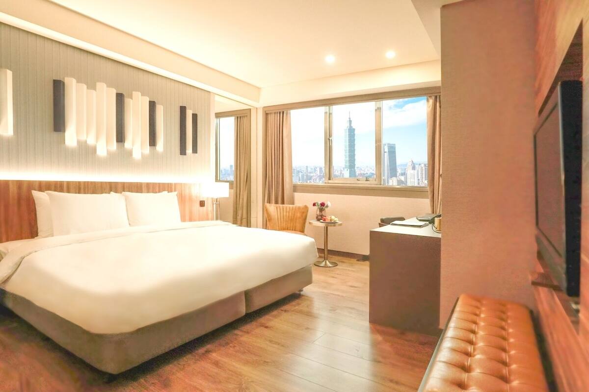 A hotel room with a large bed, a desk, a chair, a TV, and a view of the cityscape.