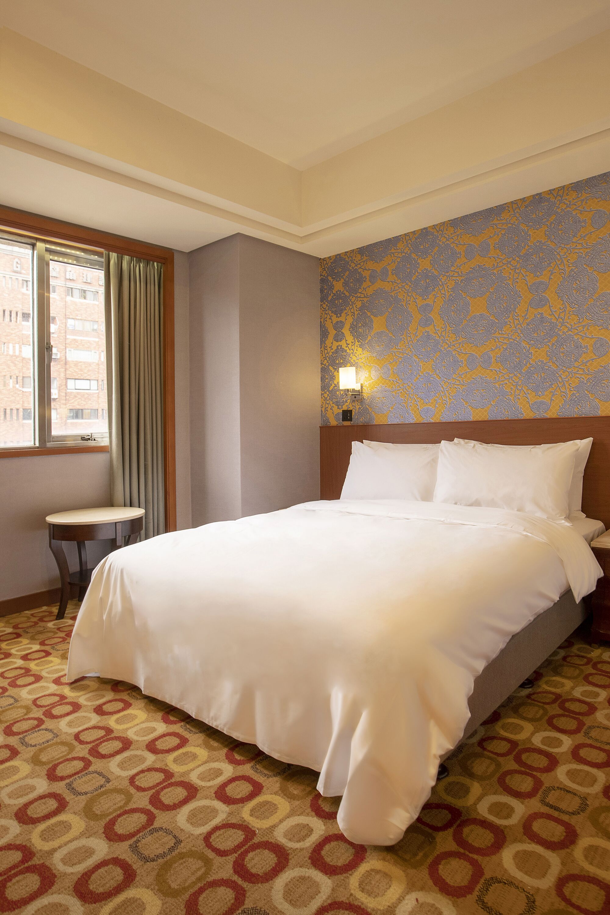 Habitación de hotel con una cama grande, una mesita pequeña, alfombra estampada, ventana con cortinas y un diseño floral en la pared.