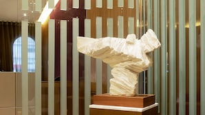 Une sculpture posée sur un piédestal en bois, dans un hall d’entrée moderne doté de cloisons en verre et d’un sol en marbre.