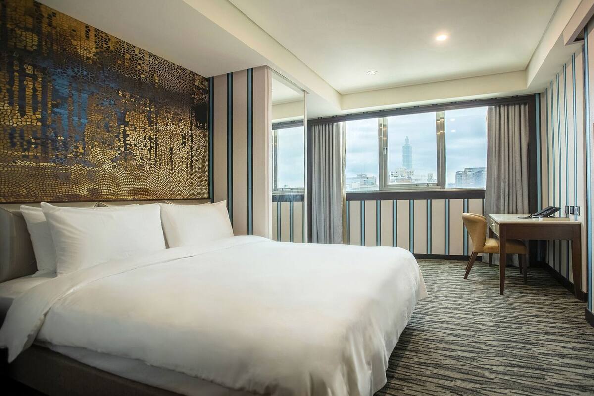 A hotel room with a large bed, a desk, a chair, and a city view.