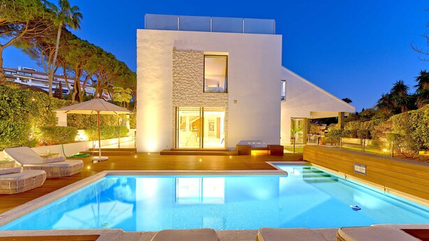 Un espace moderne aménagé au bord de la piscine, avec des chaises longues, une piscine et un bâtiment contemporain doté d’une façade en pierre et de grandes fenêtres.