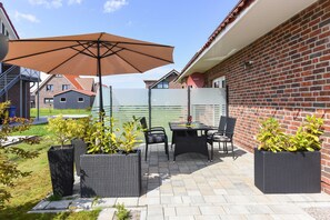 Une terrasse extérieure avec une table et des chaises, entourée de plantes en pot et d’un grand parasol.