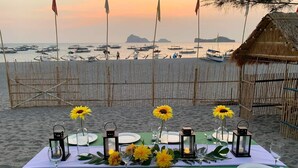 A beachside table set for a meal with a view of boats and a thatched hut in the background.