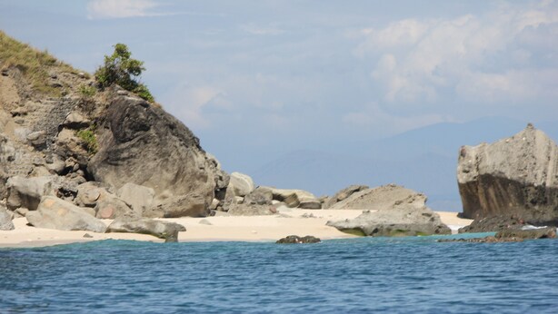 A coastal landscape with large rocks, a sandy beach, and a clear blue sky.