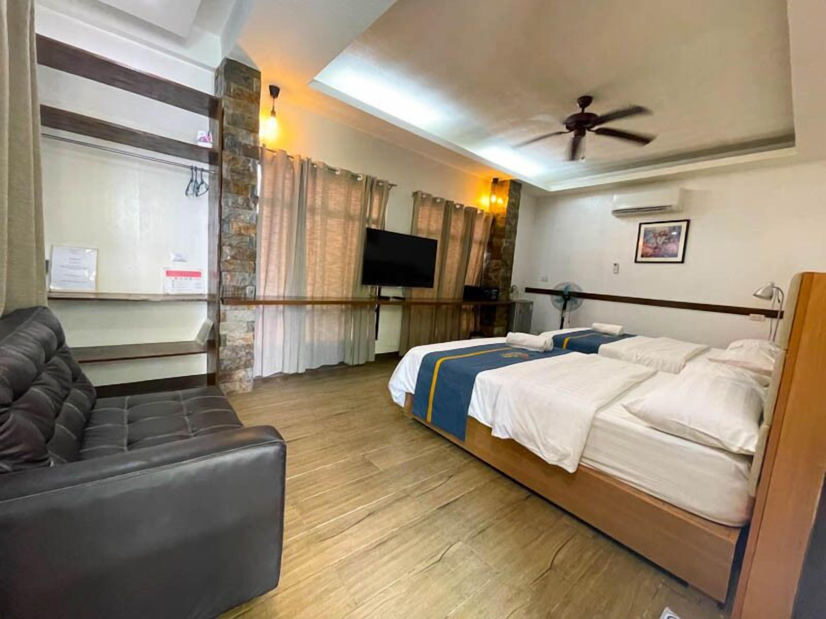 A hotel room with a sofa, two beds, a TV, and a ceiling fan.