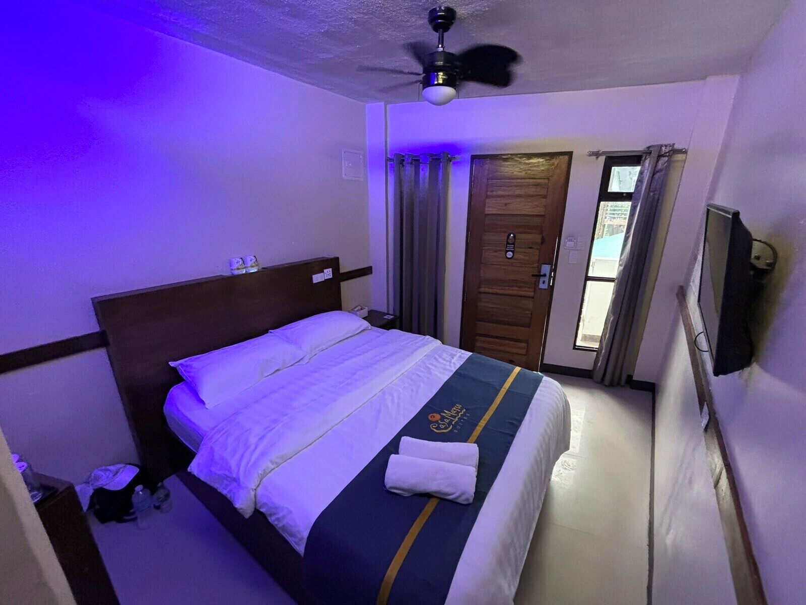 A hotel room with a bed, a ceiling fan, a wooden door, and a window with curtains.