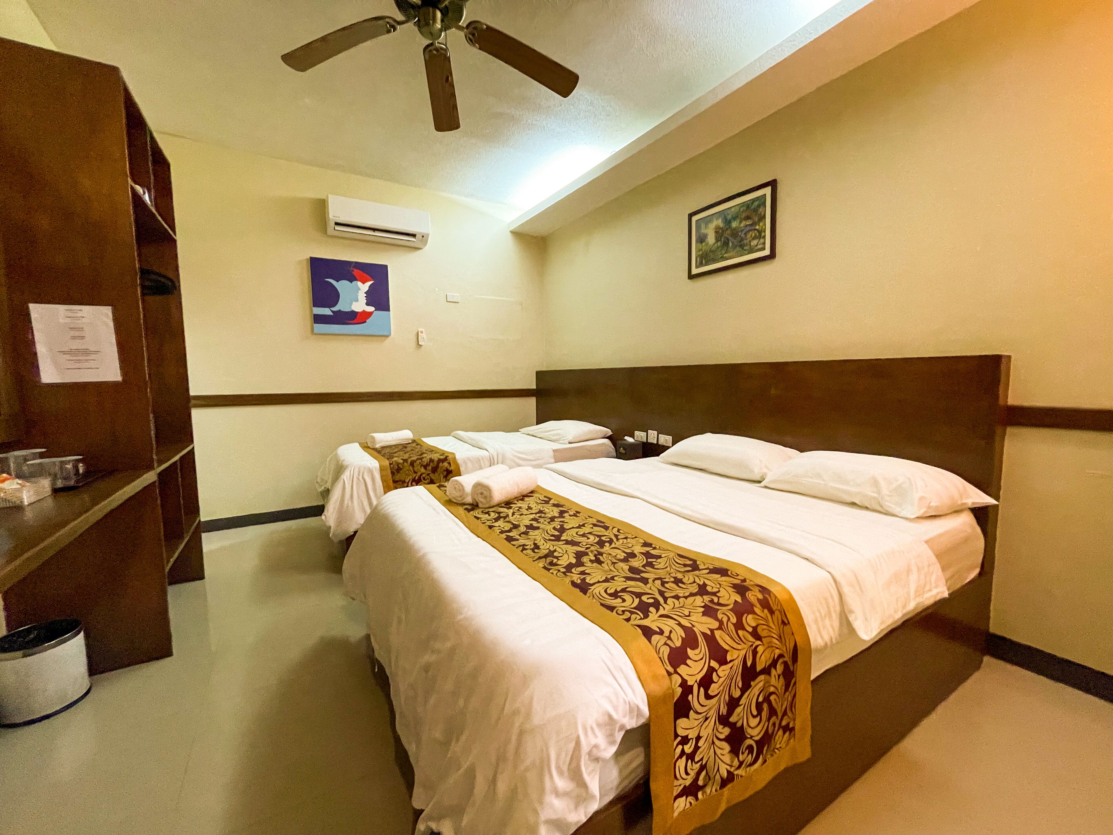 A hotel room with two beds, a ceiling fan, an air conditioning unit, a painting, and a wooden wardrobe.