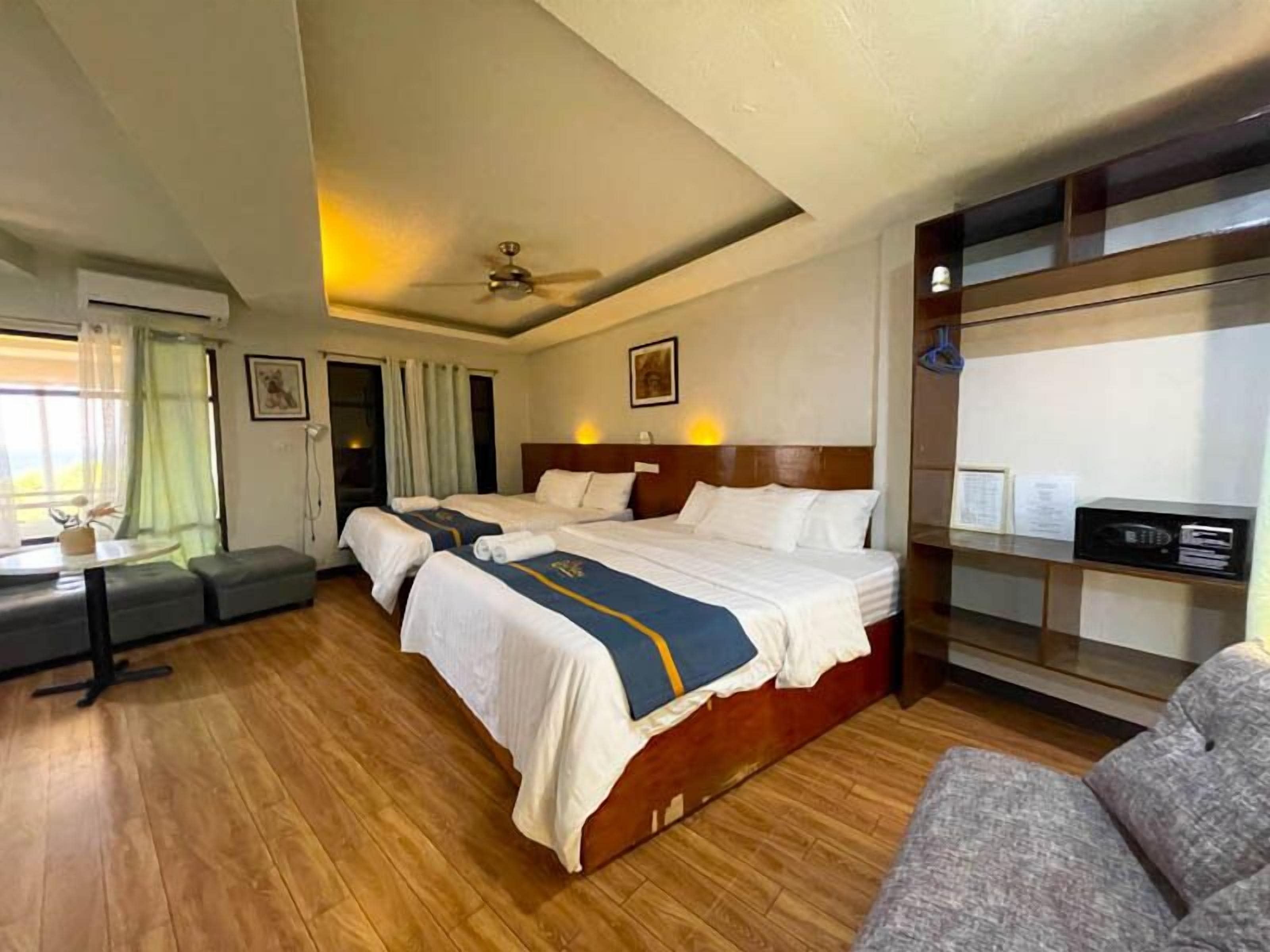 A hotel room with two beds, a desk, a sofa, and a ceiling fan.