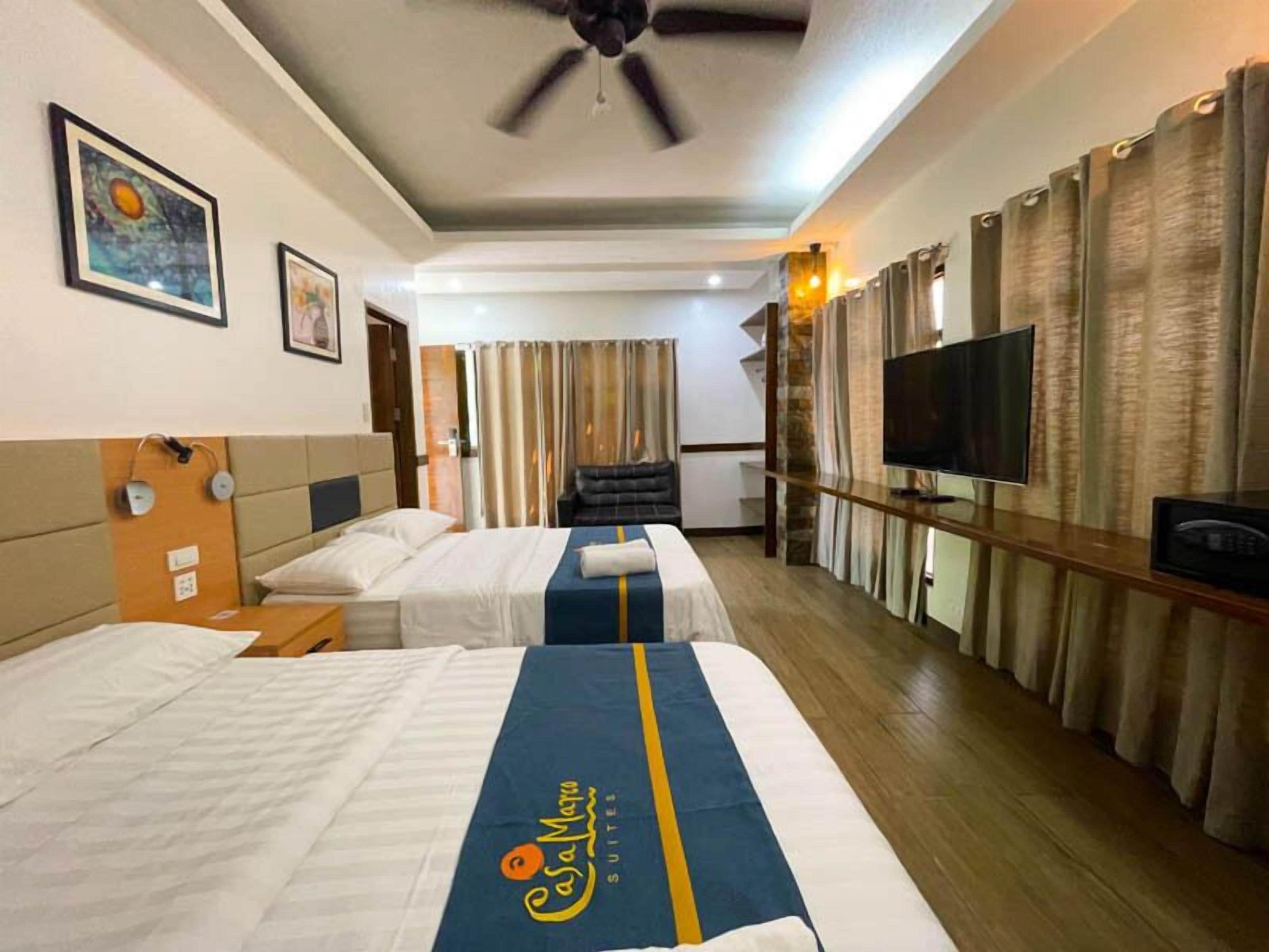 A hotel room with two beds, a TV, a ceiling fan, and a bathroom visible in the background.