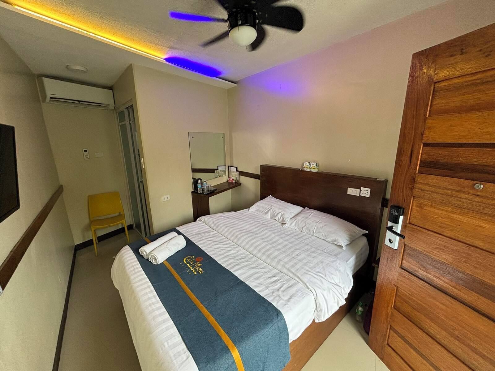 Une chambre d’hôtel avec un lit, un ventilateur de plafond, une table de chevet avec un miroir, une petite chaise et une porte en bois.