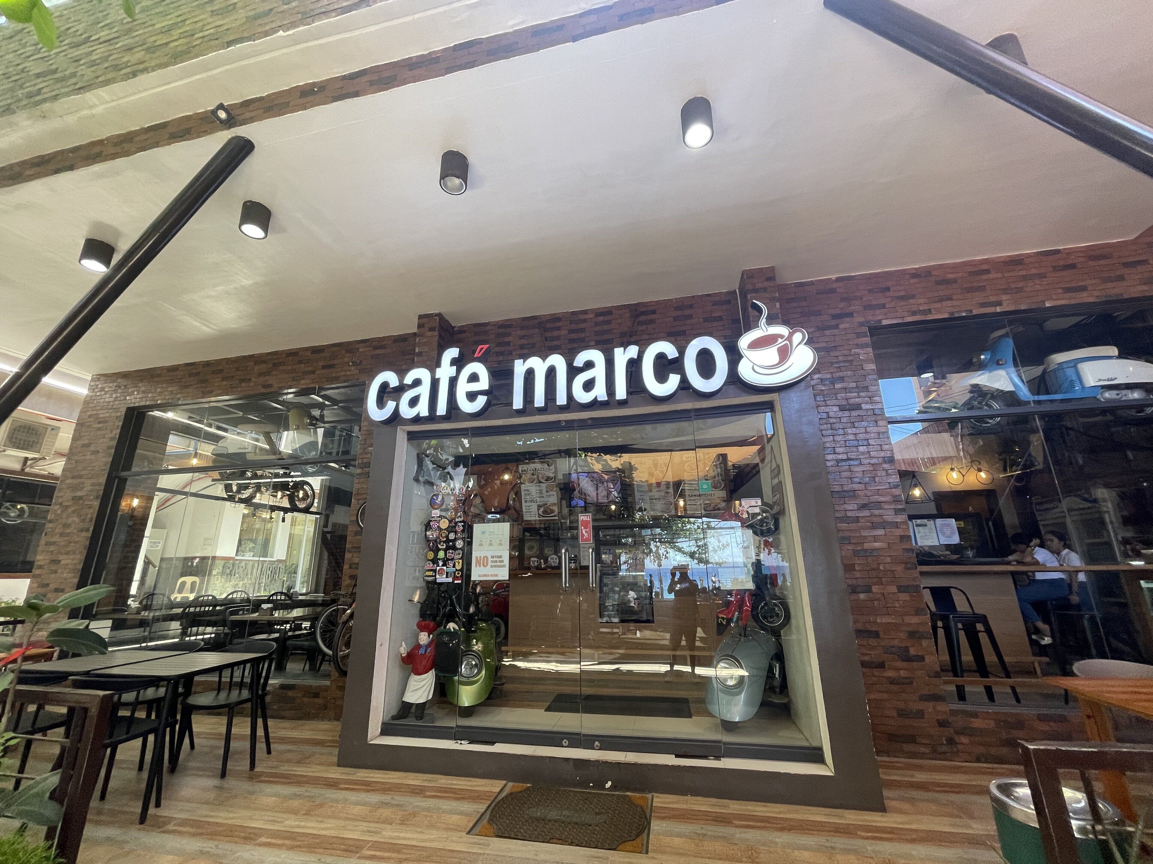 En café kaldet 'café marco' med facade i glas, udendørs siddepladser og en murstensvæg.