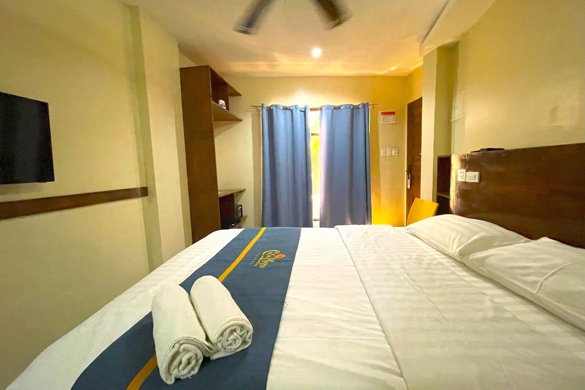 Hotellihuone, jossa on sänky, kattotuuletin, seinälle asennettu televisio ja kylpyhuone, joka näkyy oven läpi.