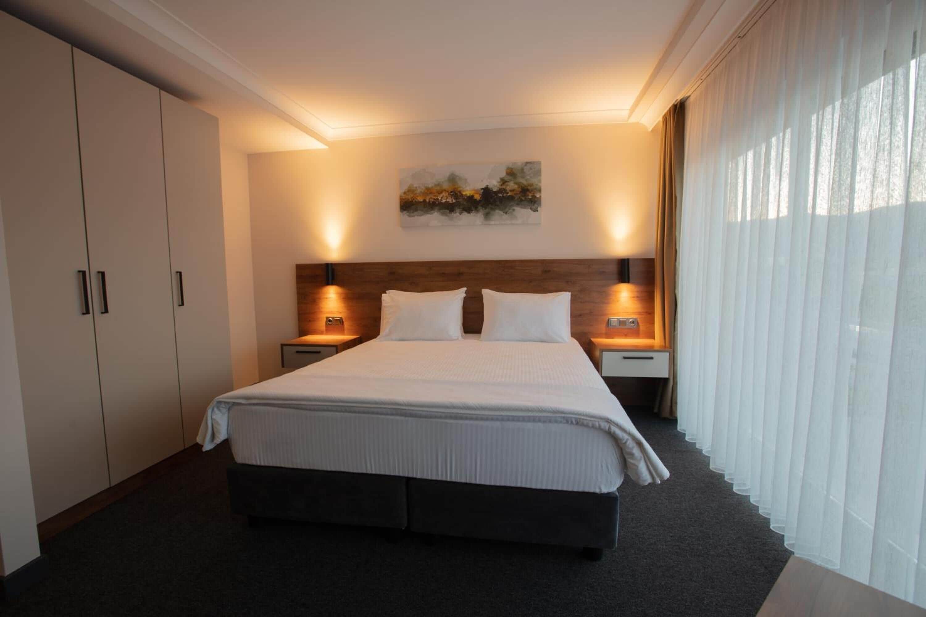 Quarto de hotel com uma cama grande, duas mesas de cabeceira, um criado-mudo e uma cabeceira de madeira, com um quadro na parede.