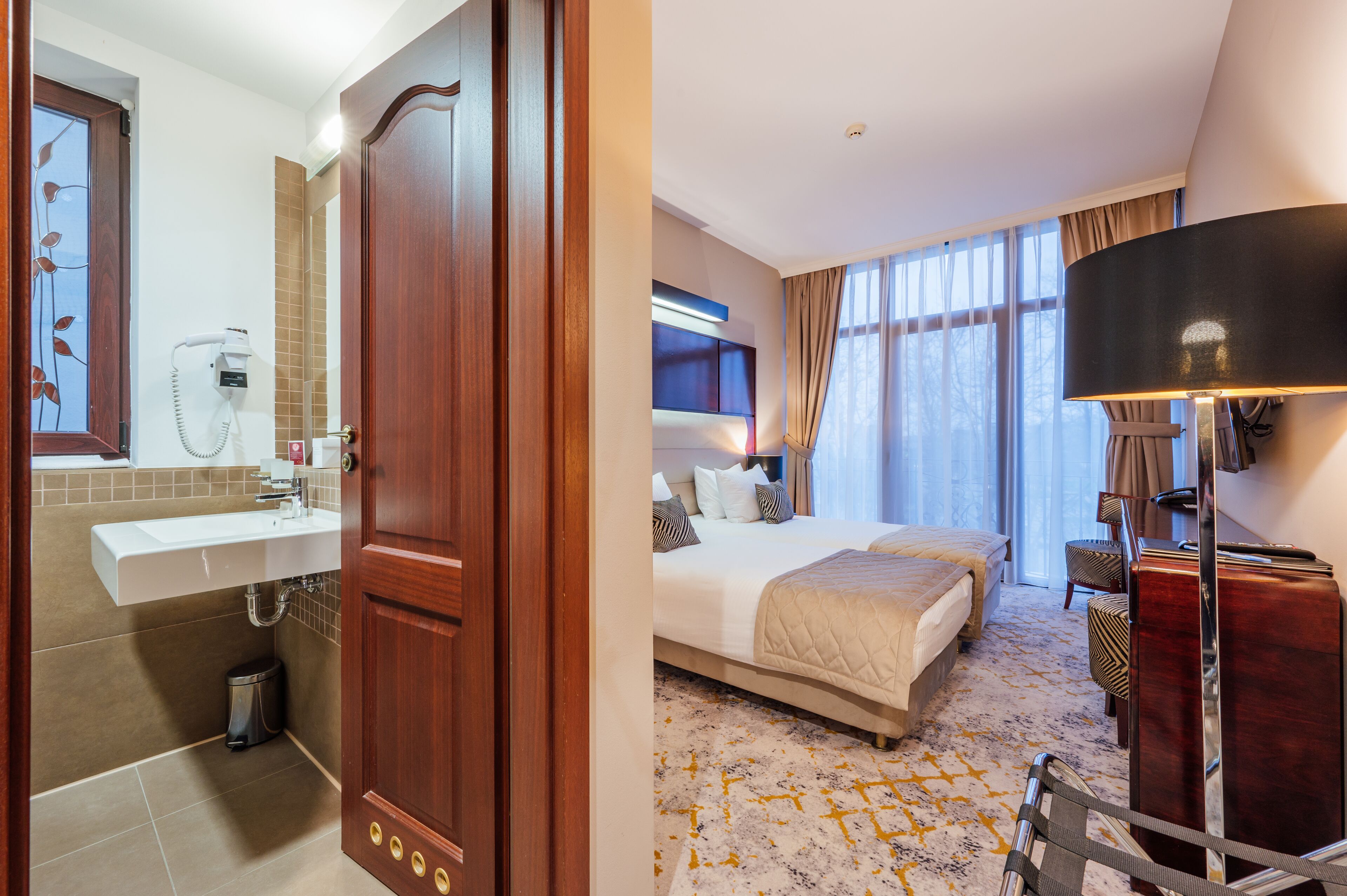 A hotel room with a wooden door, a bathroom with a sink and mirror, a bed with a headboard, and a bedside table with a lamp.