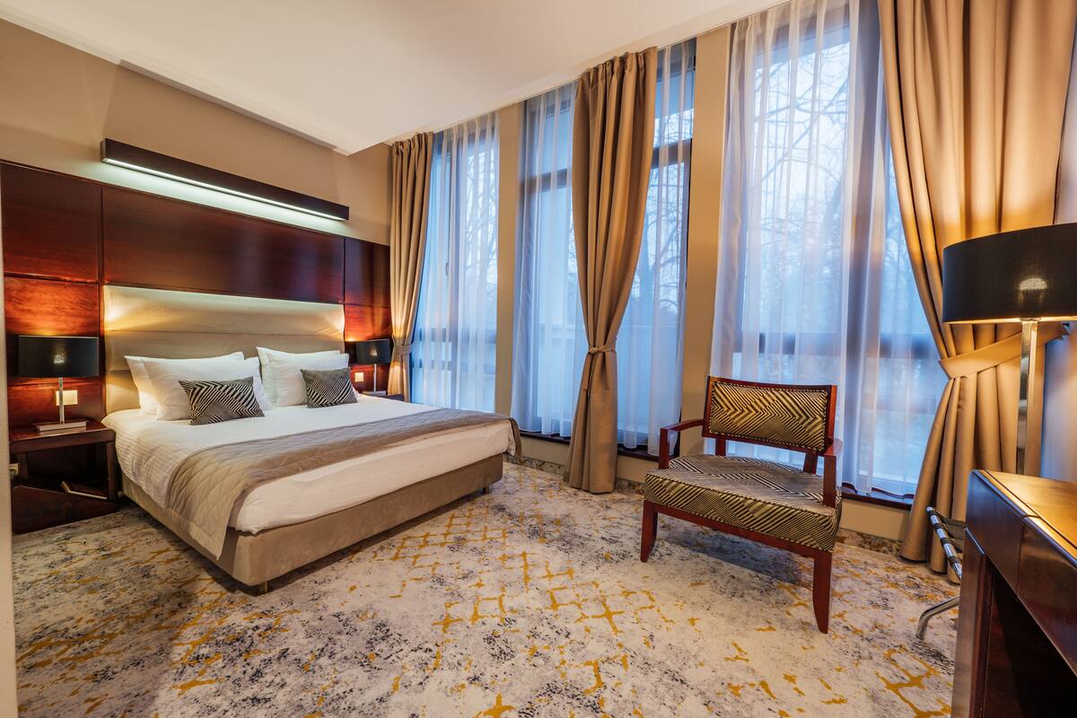 A hotel room with a large bed, two bedside tables with lamps, a chair, and a view of trees through sheer curtains.