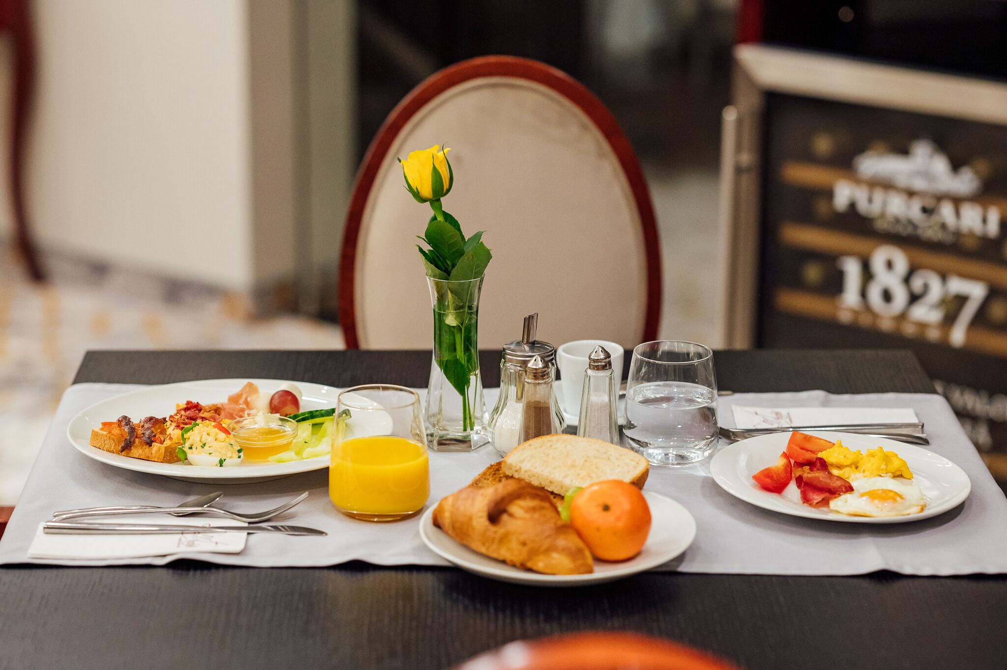 Una mesa de desayuno con platos de comida, un vaso de agua y un jarrón con una flor amarilla.