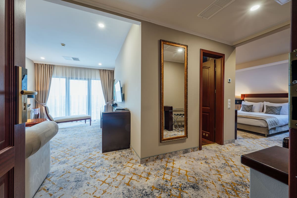 A hotel room with a large bed, a sitting area with a sofa and a desk, a large mirror, and a view of the outside through the window.