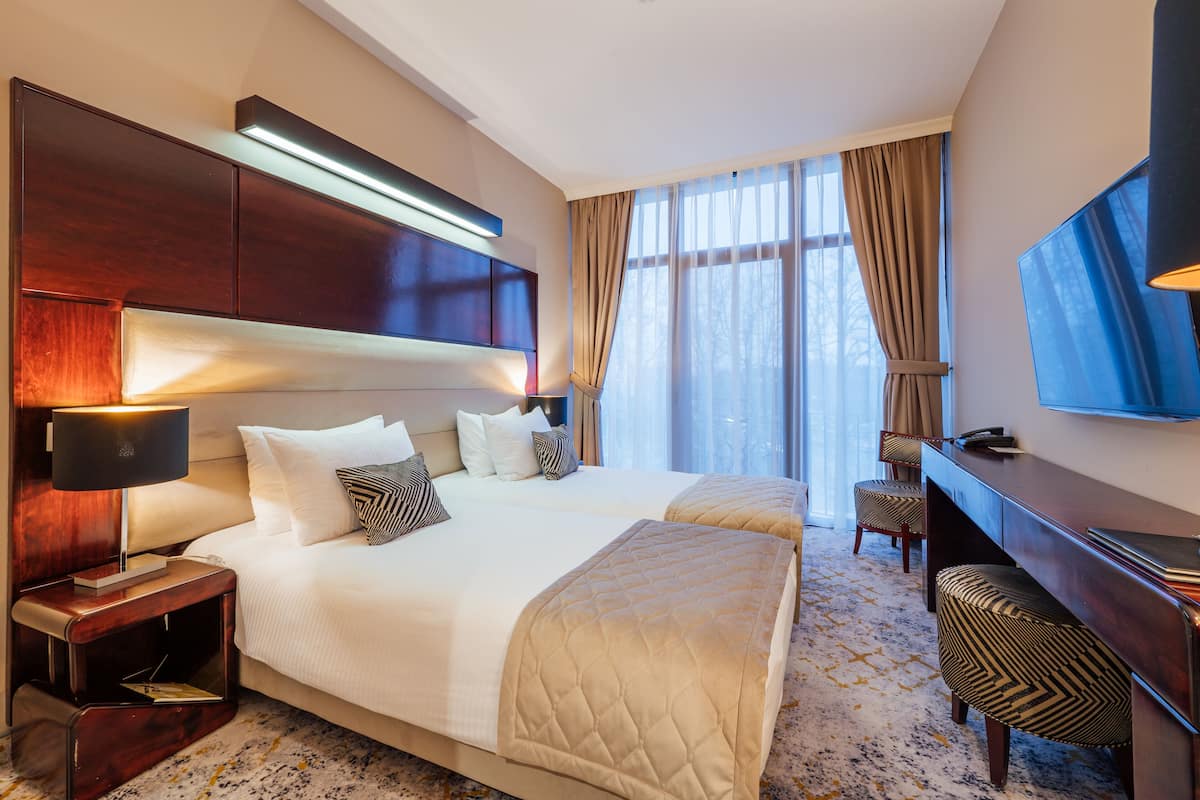 A hotel room with two beds, a desk, a chair, a TV, and a large window with curtains.