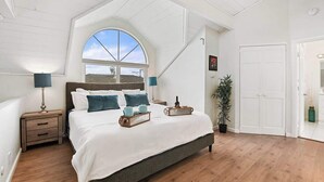 A bedroom with a large bed, a skylight, a nightstand, a lamp, a potted plant, and a door.