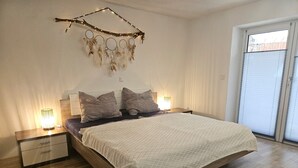 A bedroom with a bed, two bedside tables, a nightstand, a wall-mounted dreamcatcher, and a window with blinds. - Vacation apartment Alpenvorland am Lech  (Apfeldorf)
