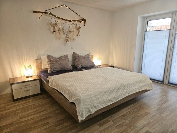 A bedroom with a bed, two bedside tables, a nightstand, a wall-mounted dreamcatcher, and a window with blinds. - Vacation apartment Alpenvorland am Lech (Apfeldorf)
