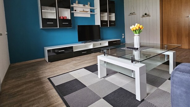 A modern living room with a blue wall, wooden beams, a glass coffee table, a TV, and a white vase with flowers. - Vacation apartment Alpenvorland am Lech  (Apfeldorf)