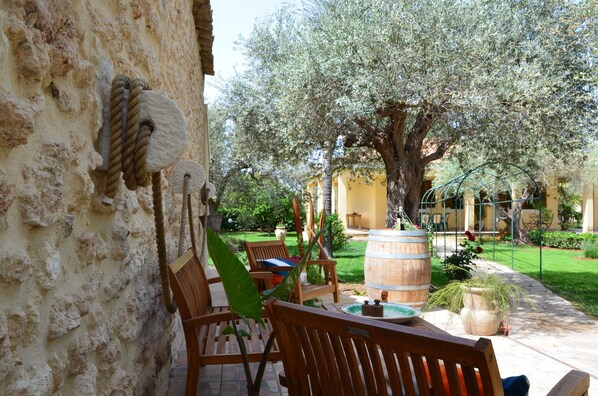 A courtyard with stone walls, wooden furniture, a large tree, and a barrel.
