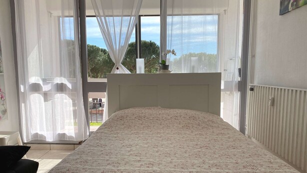 A bedroom with a bed, a window with sheer curtains, a radiator, and a view of the outside. - Studio 1st floor with wifi, park view (Balaruc-les-Bains)