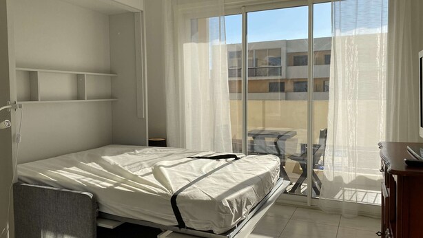 Une chambre à coucher comprenant un lit, une petite table, une chaise, une armoire et une porte-fenêtre coulissante donnant sur un balcon.