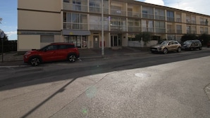 A multi-story building with a flat facade, a ground-level entrance, and a parking area with several cars. - Cozy Studio Near Thermal Baths with Balcony, A/C, and Modern Amenities (Balaruc-les-Bains)