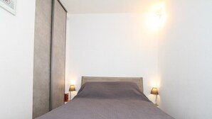 A single bed with a grey bedspread, two bedside lamps, and a wooden headboard in a room with white walls. - Studio in Balaruc-les-Bains with AC and Wifi (Balaruc-les-Bains)