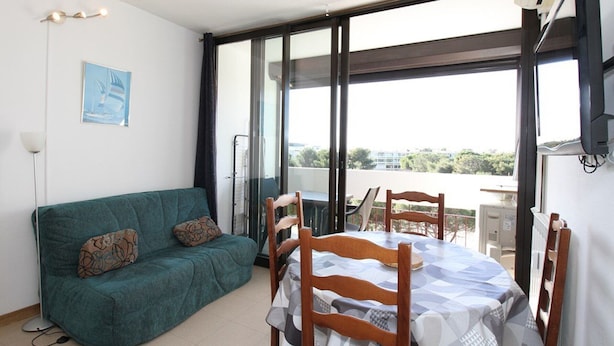 A room with a teal sofa, a wooden dining table with chairs, a large window with a view, and an air conditioning unit. - Studio in Balaruc-les-Bains with AC and Wifi (Balaruc-les-Bains)