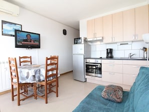 A compact kitchen with a dining area, a sofa, and a TV displaying a news program.