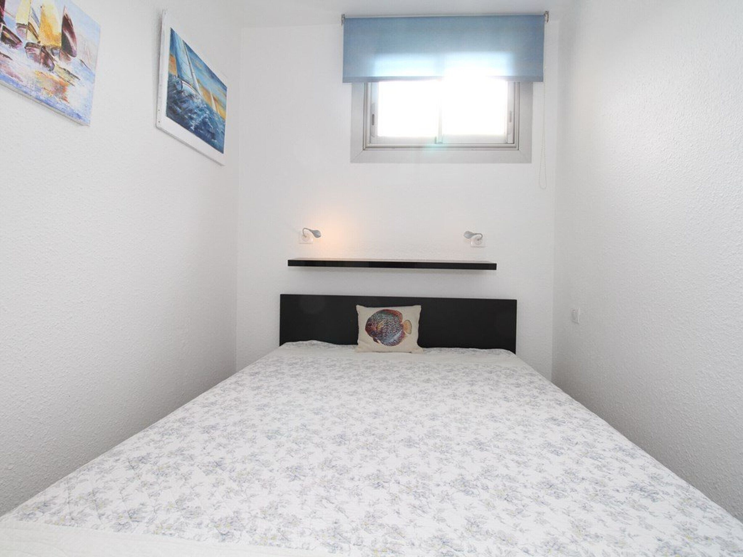 Un dormitorio con una cama, una ventana con persiana azul y dos cuadros en la pared.