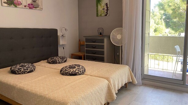 Ein Schlafzimmer mit einem Bett, Kissen, einem Nachttisch, einer Lampe, einem Ventilator, einem Schminktisch und einem Fenster mit Blick auf Bäume.
