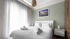 Un letto rifatto con lenzuola bianche e due asciugamani arrotolati, un comodino con una lampada e un quadro raffigurante un paesaggio urbano.