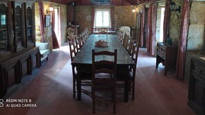 A spacious dining hall with a long wooden table and chairs, stone walls, and a high wooden ceiling with exposed beams.