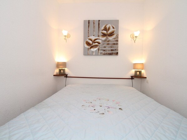 A bedroom with a bed, two wall-mounted lamps, and a floral artwork on the wall. - Charming T1 Apartment with A/C, 100m from Thermal Baths, Fully Equipped Kitchen, Private Loggia (Balaruc-les-Bains)