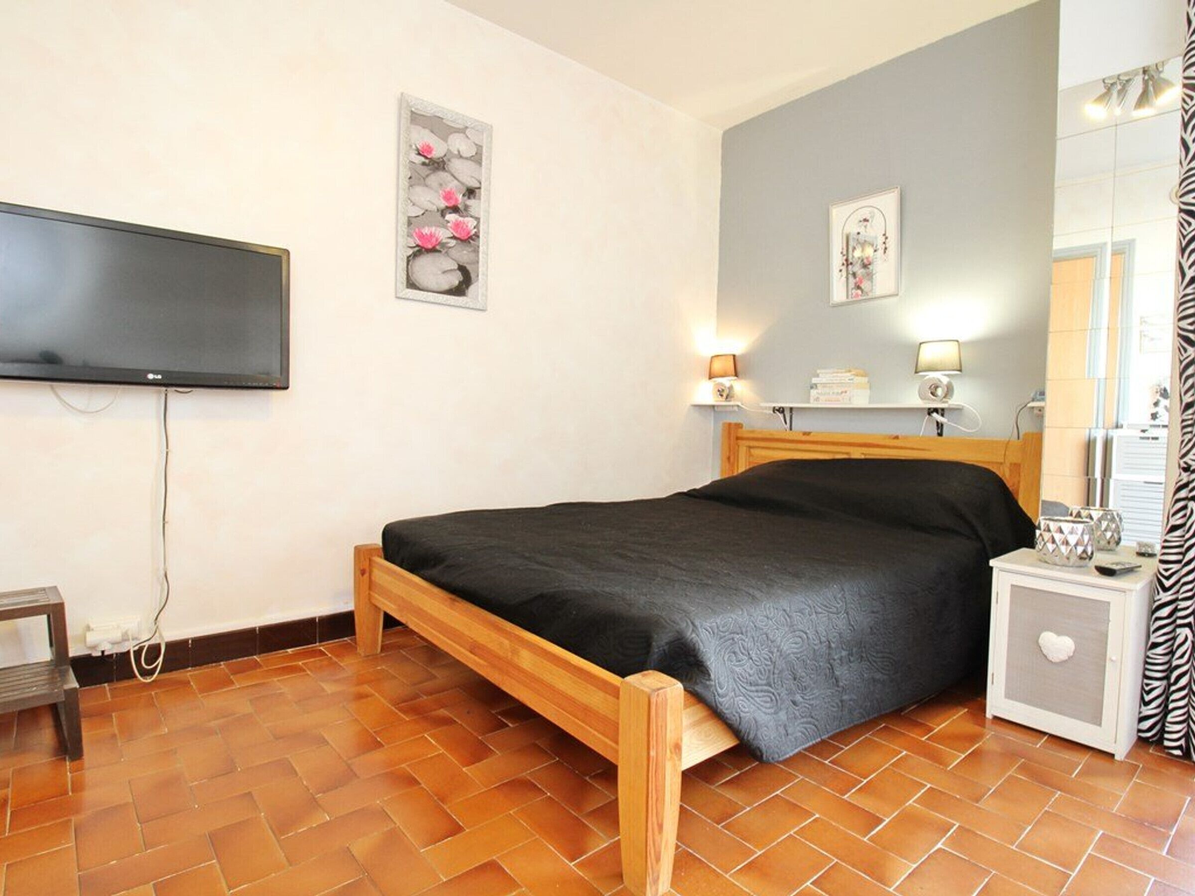 Un dormitorio con cama de madera, televisor de pantalla plana montado en la pared, una mesita de noche con lámpara y un espejo.