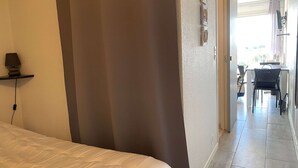Et hotelværelse med en seng, et skrivebord og en stol. Der er et gardin ved vinduet, og en lampe på en hylde.
