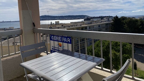 En balkon med et bord og stole, der kigger ud over et kystområde med bygninger og vand.